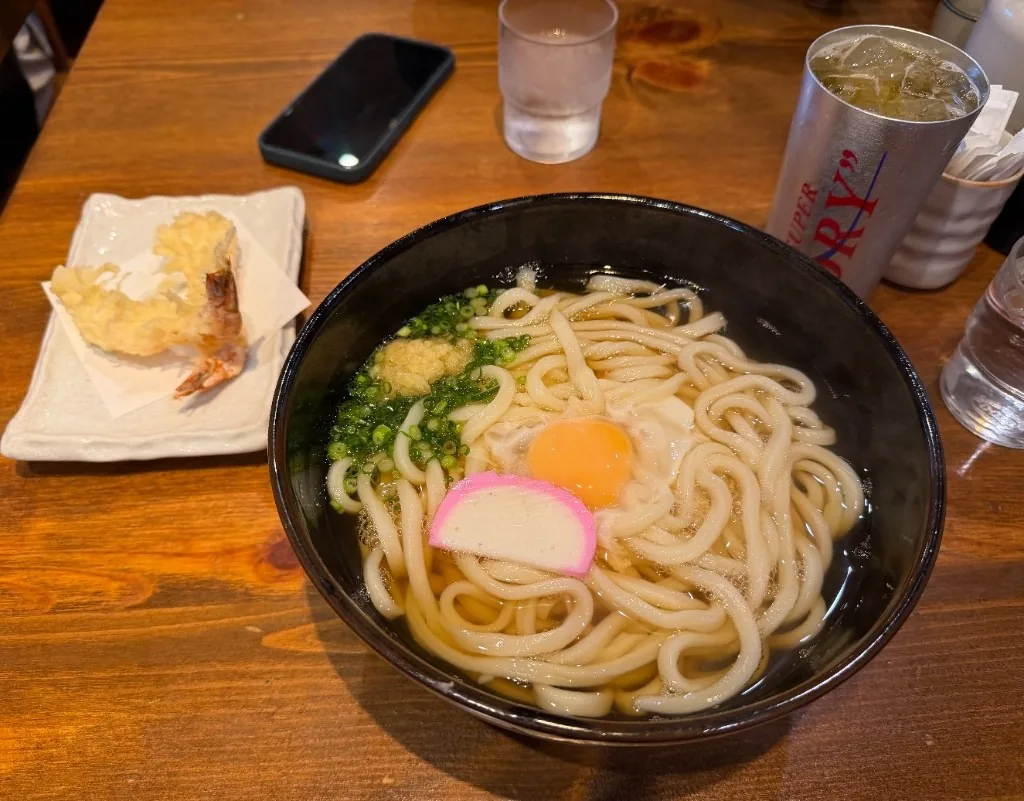 A bowl of Sanuki udon in clear dashi with a raw egg yolk, kamaboko, ginger and scallions, with shrimp tempura on the side